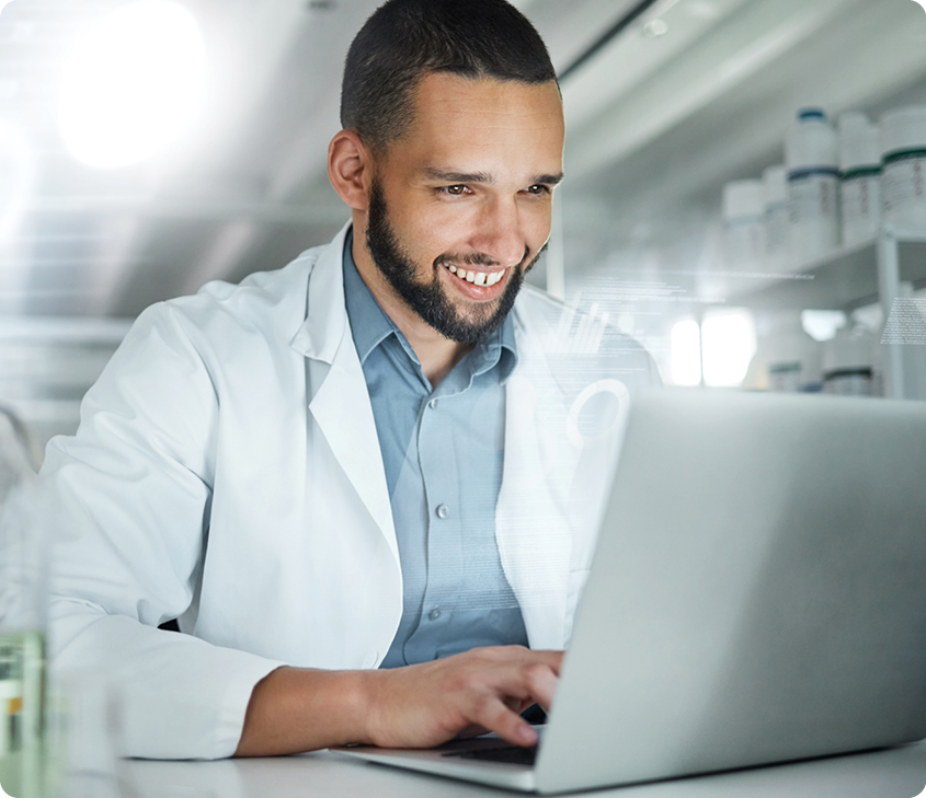 smiling male in white lab coat sitting in front of a laptop